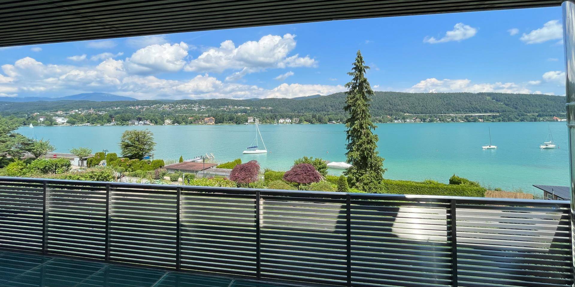 Panoramablick vom Balkon auf den glitzernden See und die umliegende Natur unter strahlend blauem Himmel.