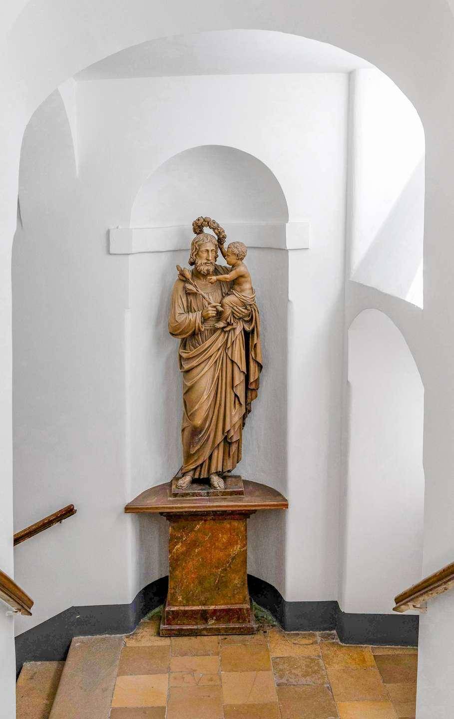 Eine klassische Holzstatue auf einem Podest in einem hellen Treppenhausbereich.