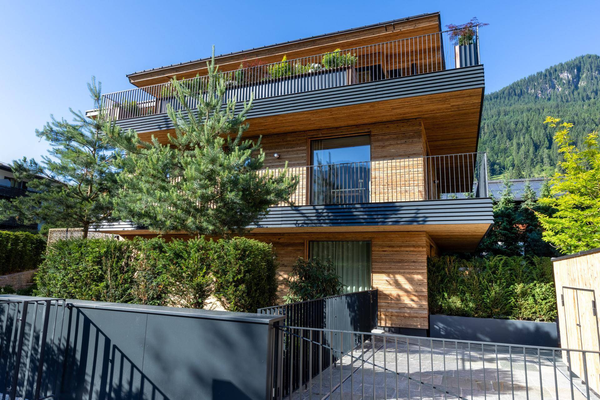 Moderne Architektur mit Holzfassade, mehreren Balkonen und gepflegter Bepflanzung in alpiner Umgebung.