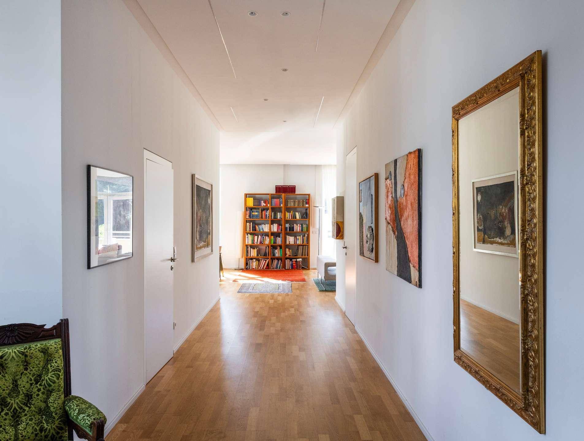 Langer, heller Flur mit Holzboden, Kunstwerken an den Wänden und Bücherregal am Ende.