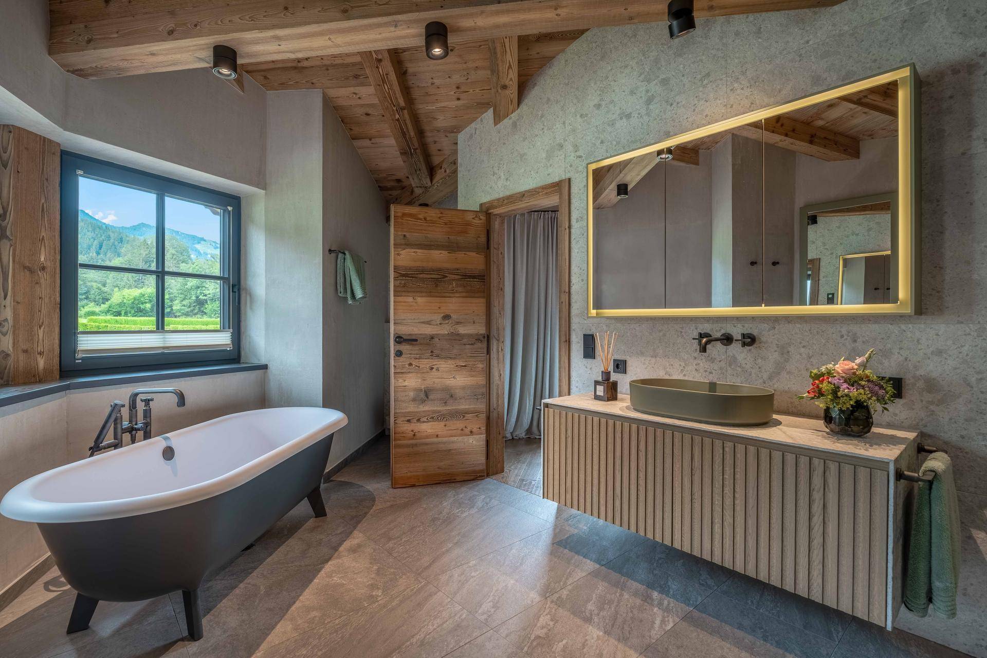 Luxuriöses Badezimmer mit freistehender Badewanne, Holzdecke und Fenster mit Grünblick.
