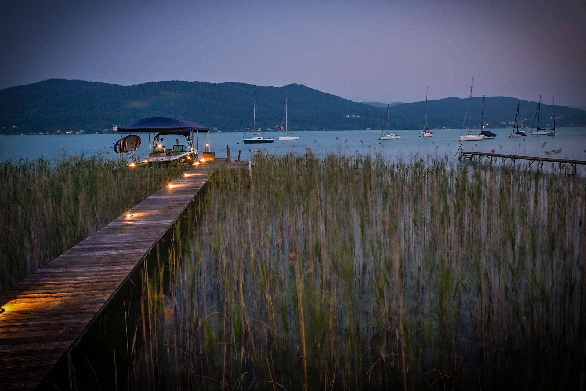 Beleuchteter Holzsteg führt zu einem Bootsanleger am See, umgeben von Schilf und Bergen.