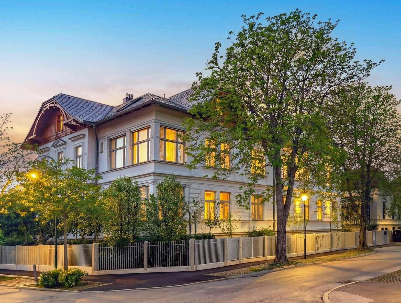 Elegantes Gebäude mit klassischer Architektur, umgeben von Bäumen und einem gepflegten Zaun bei Sonnenuntergang.