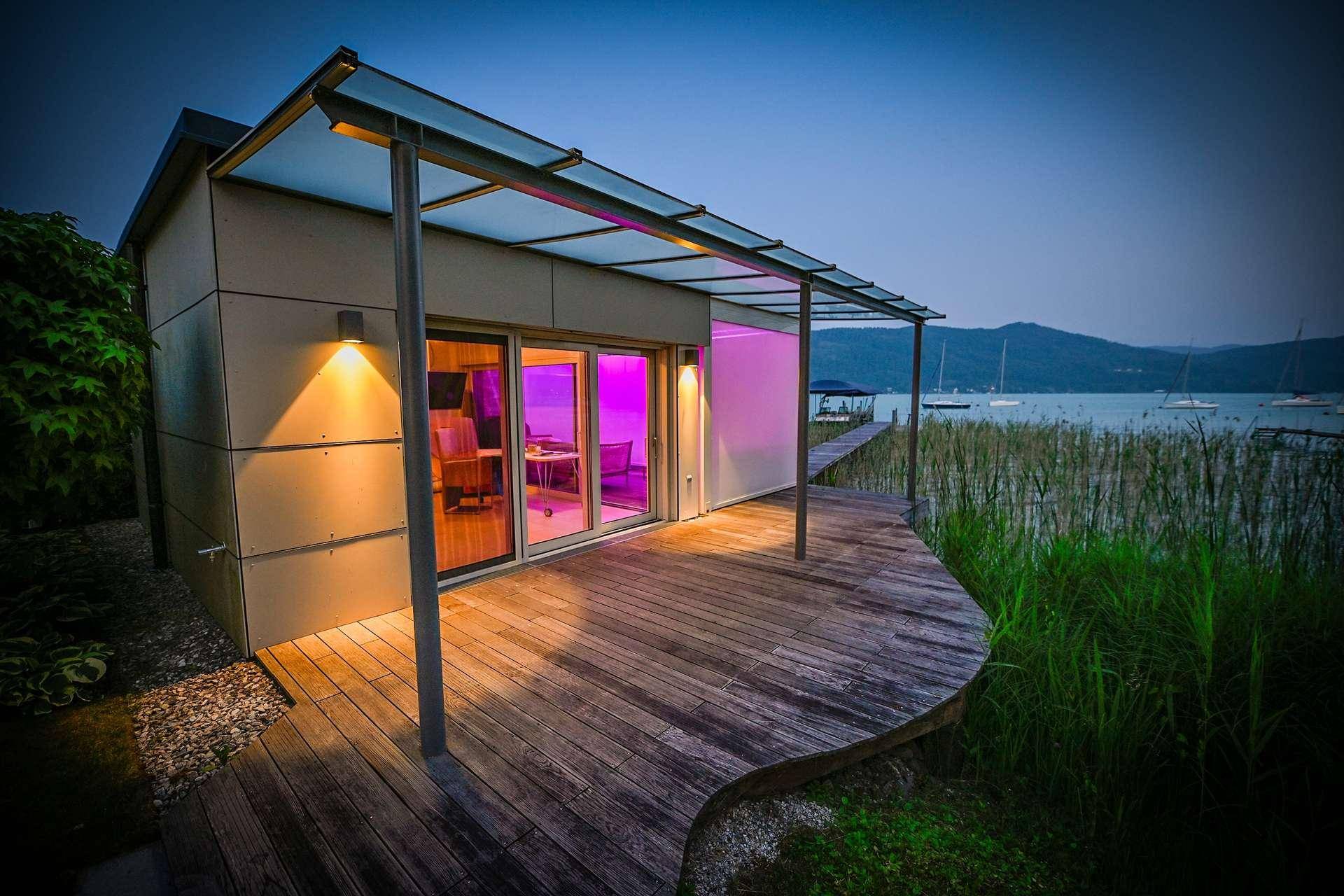 Modernes Gebäude mit Holzterrasse direkt am See, umgeben von Schilf und mit Bergblick.