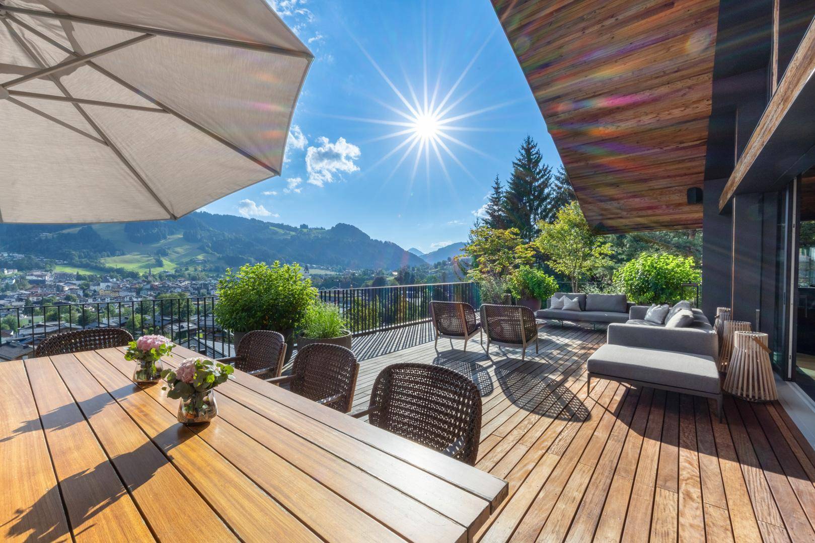 Sonnige Holzterrasse mit Esstisch und Lounge-Möbeln, die einen weiten Blick auf die Berge und die Stadt bietet.