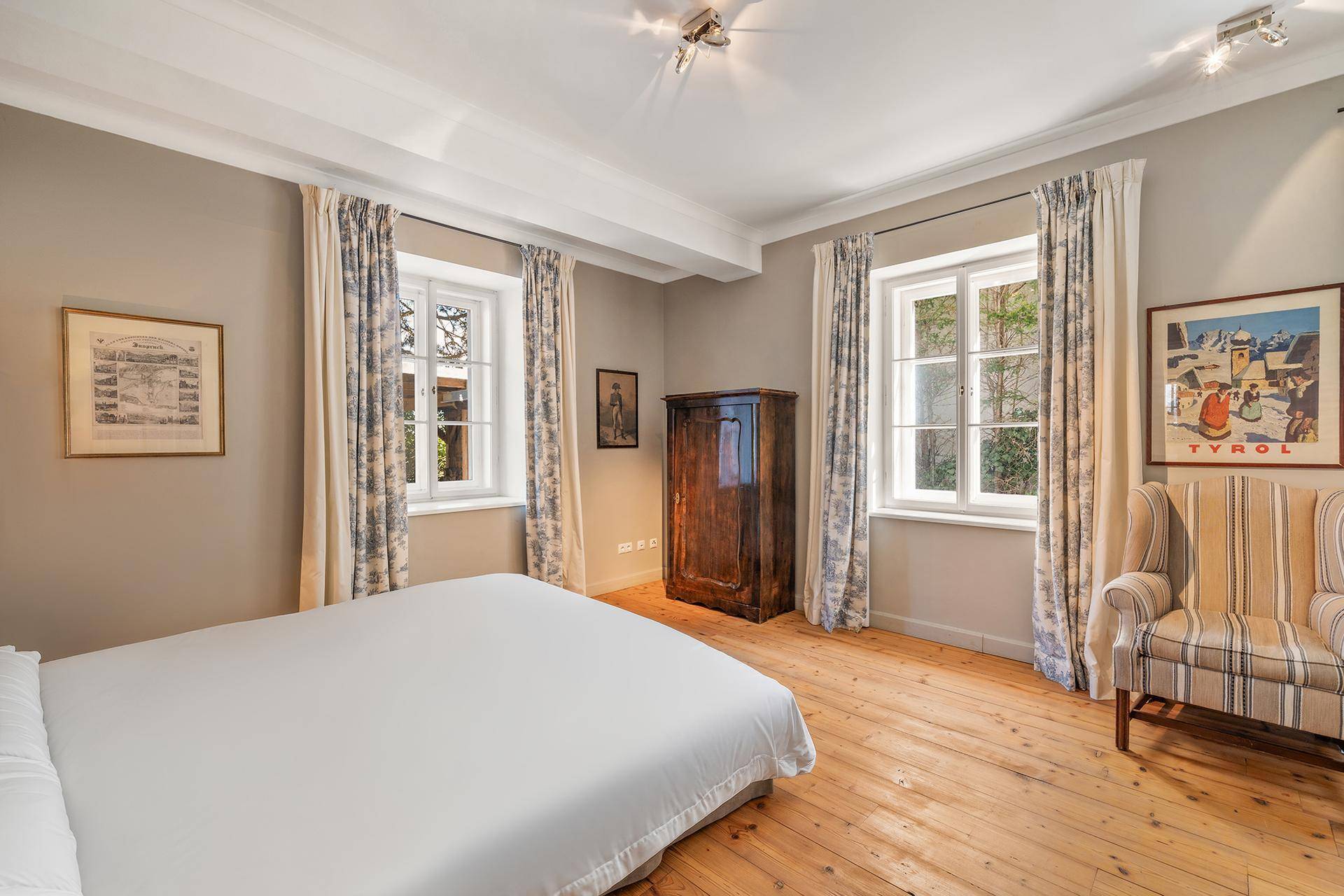 Helles Schlafzimmer mit Holzboden, zwei Fenstern und klassischem Mobiliar.
