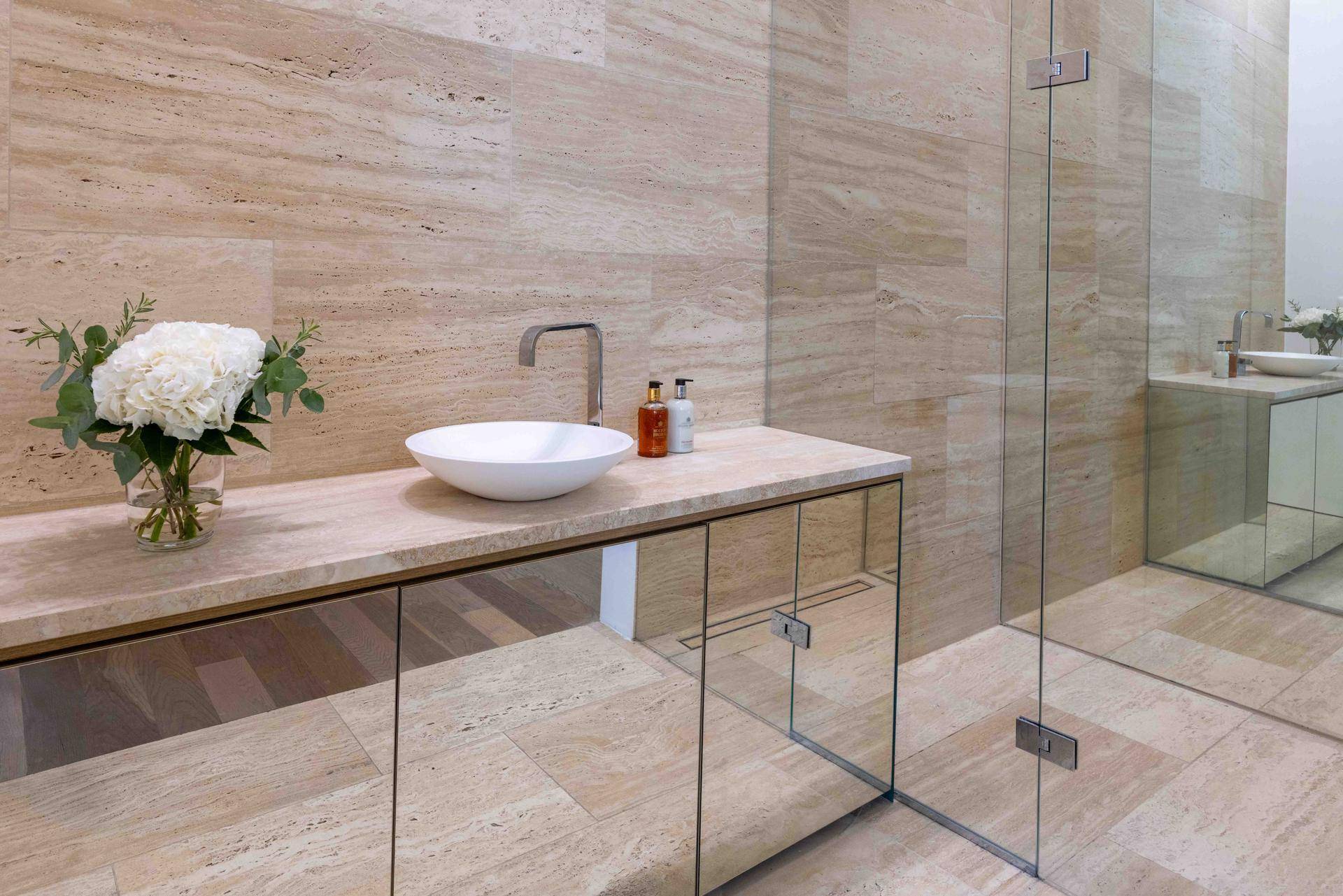 Modernes Badezimmer mit Natursteinfliesen, einem eleganten Einzelwaschbecken und verspiegelten Unterschränken.