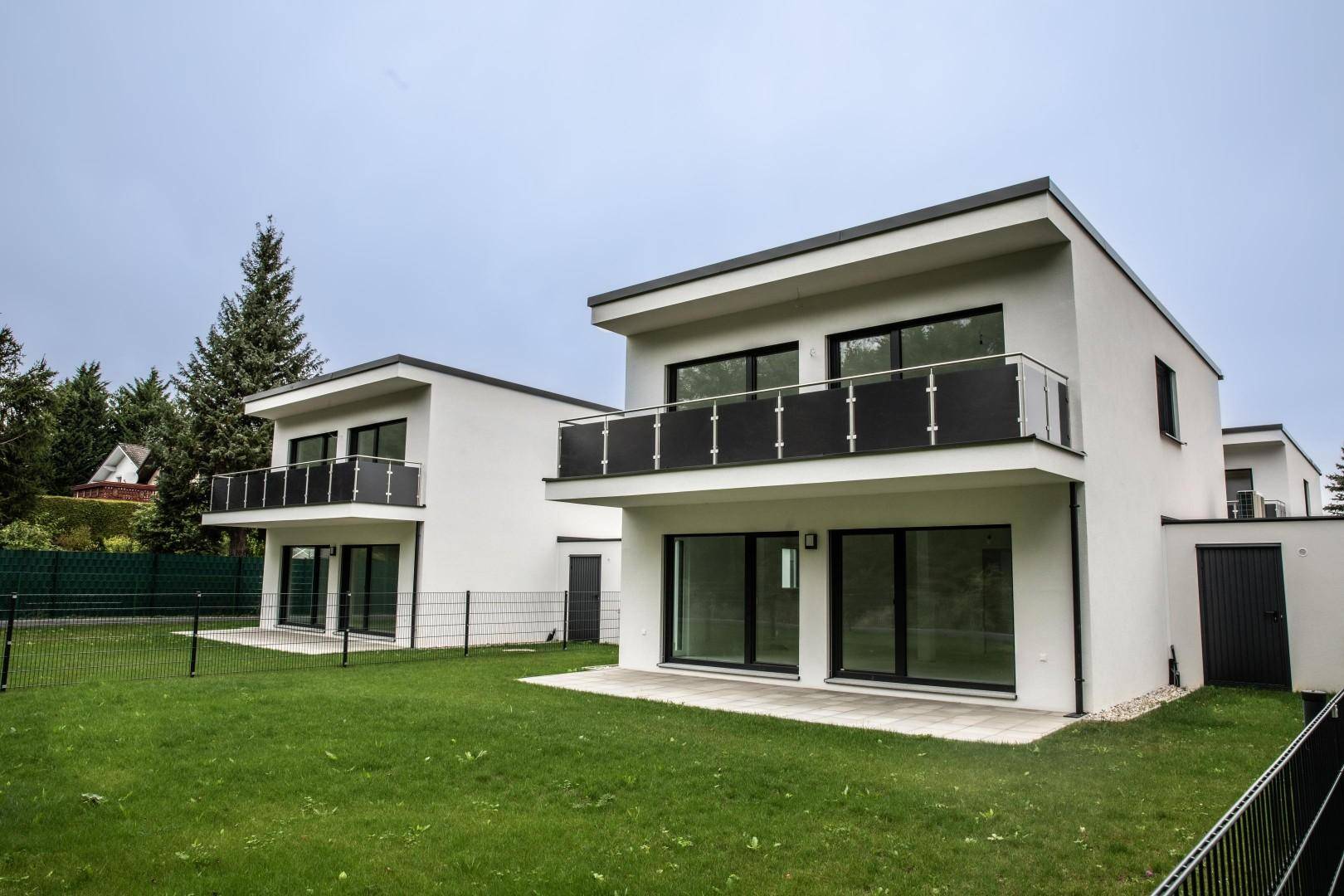 Zwei moderne Neubauhäuser mit weißen Fassaden, Balkonen und grünen Gärten.
