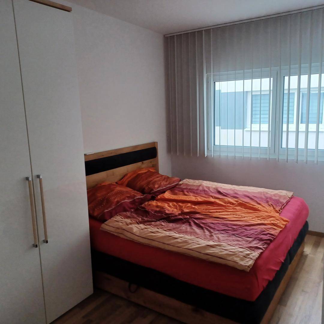 Helles Schlafzimmer mit Doppelbett, Holzfußboden und Fenster mit vertikalen Jalousien.