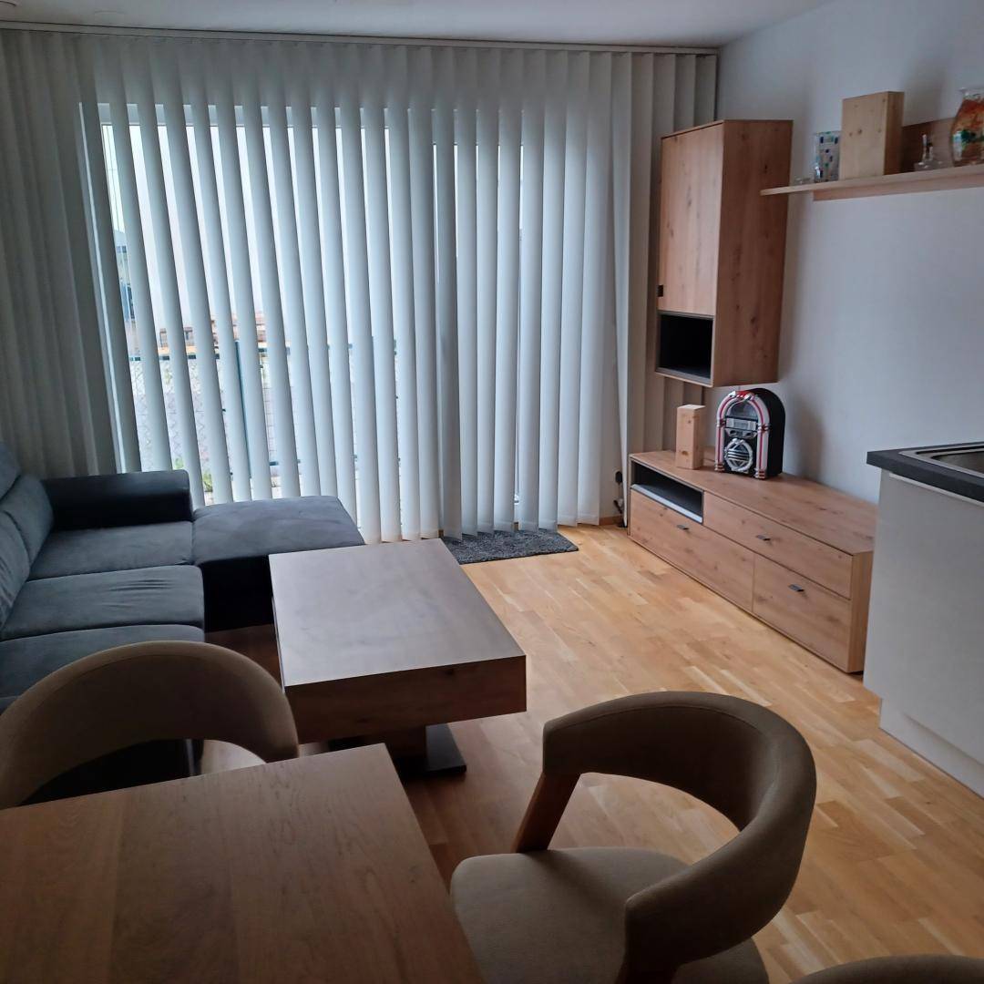 Gemütliches Wohnzimmer mit Ecksofa, Holzboden und vertikalen Jalousien am Fenster.