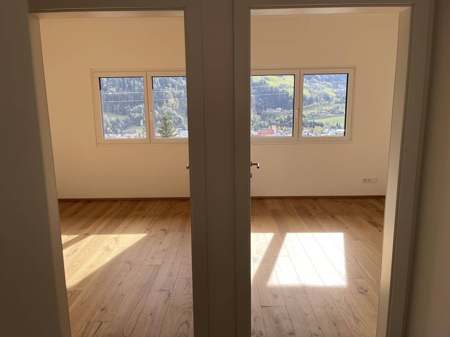 Blick durch eine Tür in zwei helle, leere Räume mit Holzboden und Fenstern mit Bergblick.