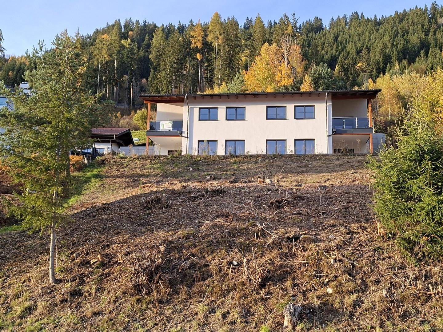 Modernes weißes Haus auf einem Hanggrundstück mit herbstlicher Waldkulisse im Hintergrund.
