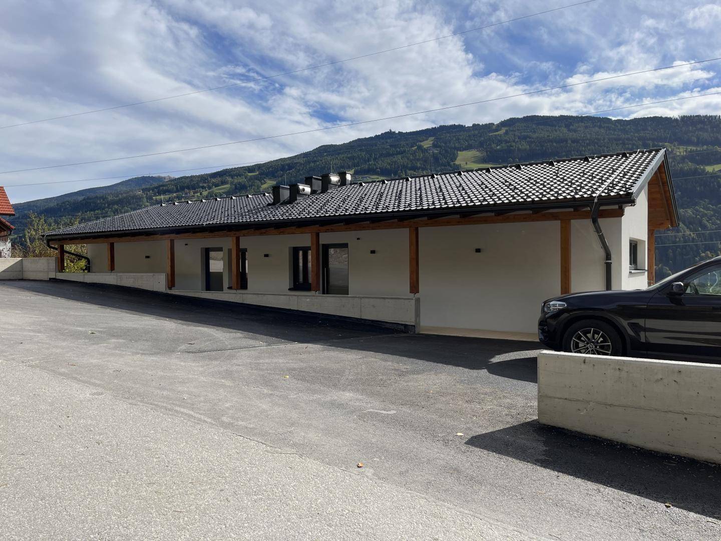 Modernes Gebäude mit dunklem Ziegeldach, weißer Fassade und langer gepflasterter Auffahrt vor Bergkulisse.