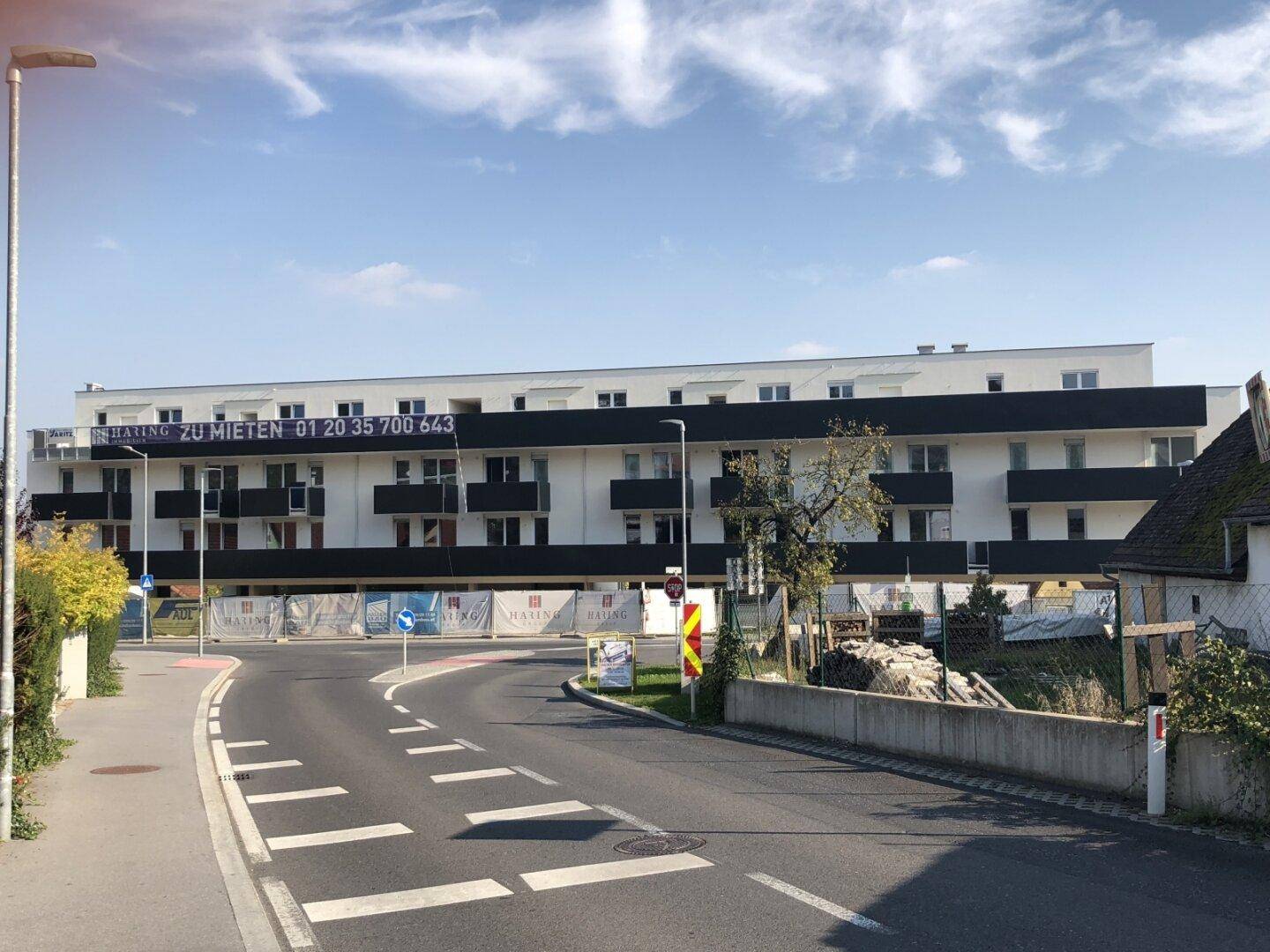 Modernes Neubauwohngebäude an einer Straße mit Werbebannern und Baustellenabsperrungen.