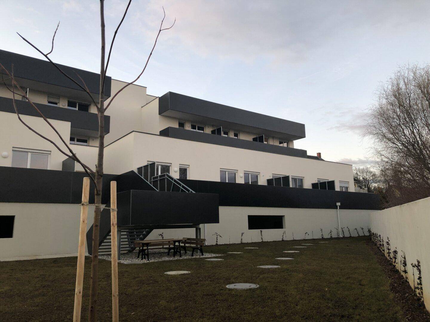 Gepflegte Rasenfläche hinter einem modernen Wohngebäude mit Außentreppe und Sitzgelegenheiten.