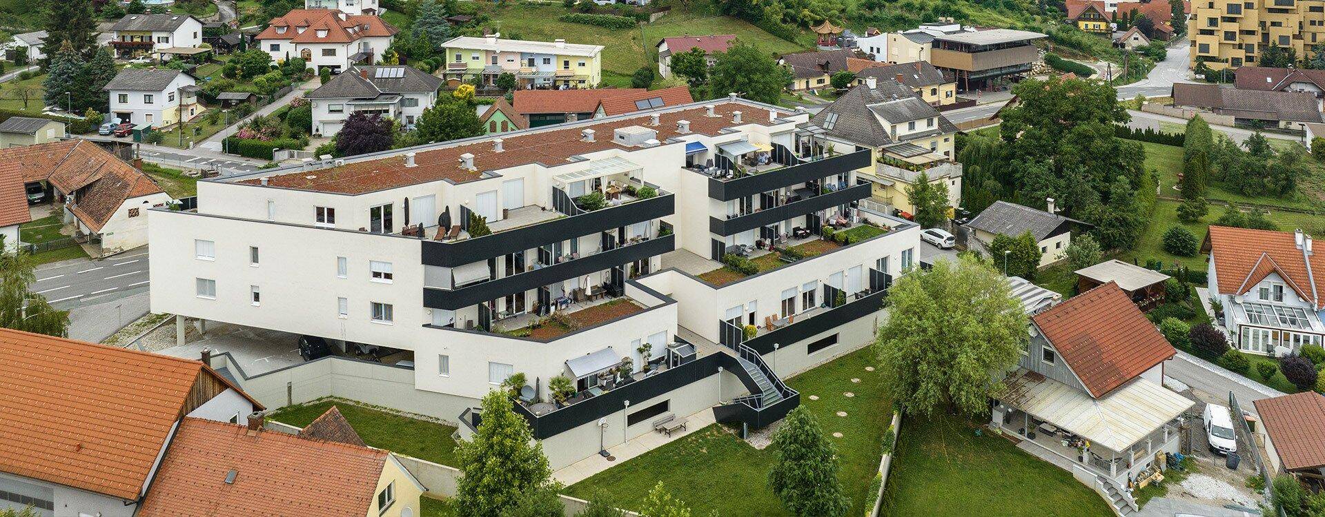 Luftaufnahme eines modernen Wohngebäudes mit Terrassen und Balkonen, umgeben von Grünflächen und Häusern.