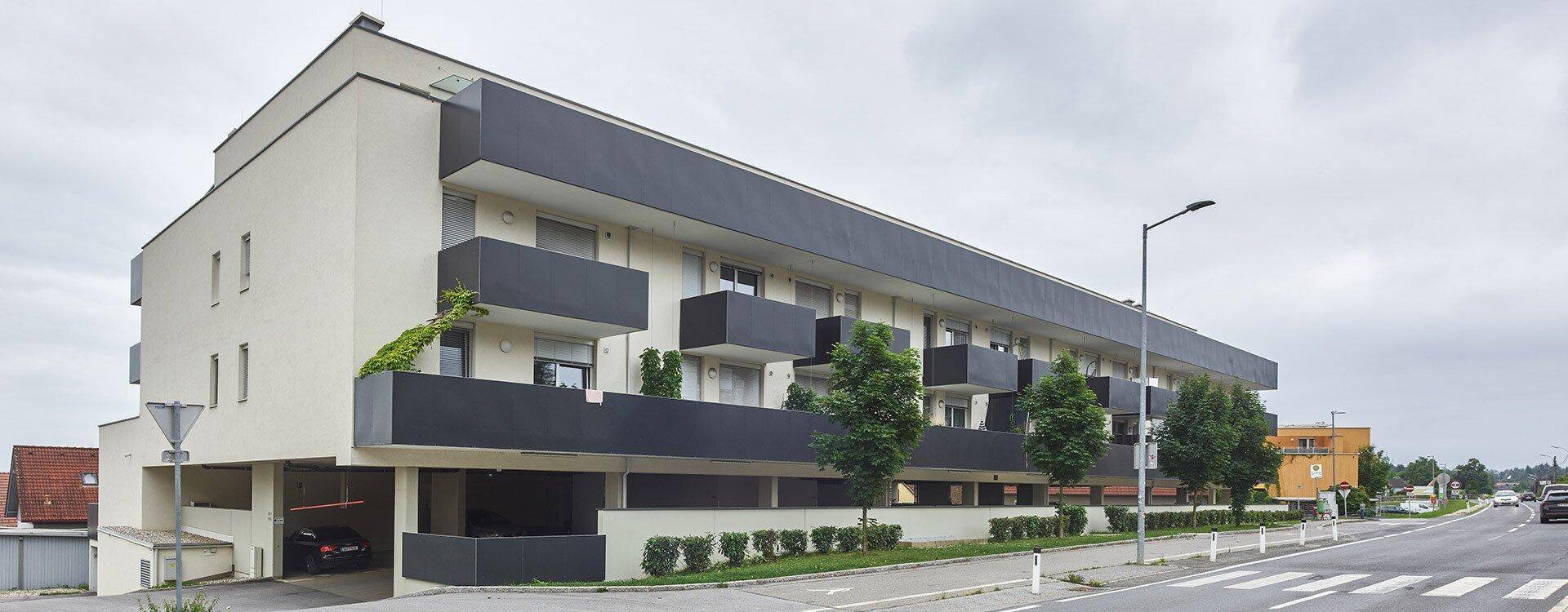 Langes, modernes Wohngebäude mit dunklen Balkonen und integrierter Tiefgarage an einer Straße.