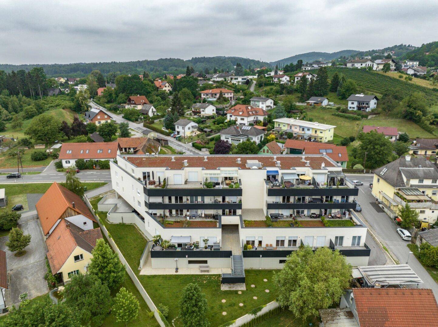 Luftaufnahme eines modernen Wohngebäudes mit Terrassen und Balkonen in hügeliger Landschaft.