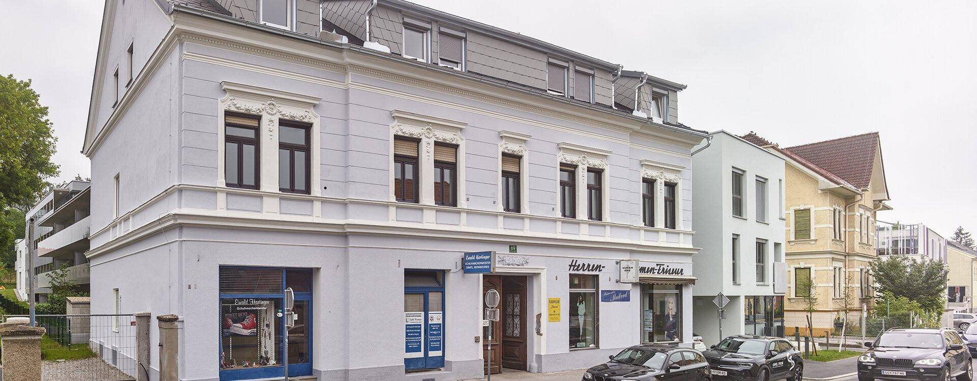 Historisches Gebäude mit Geschäften im Erdgeschoss und modernen Anbauten an einer belebten Straße.
