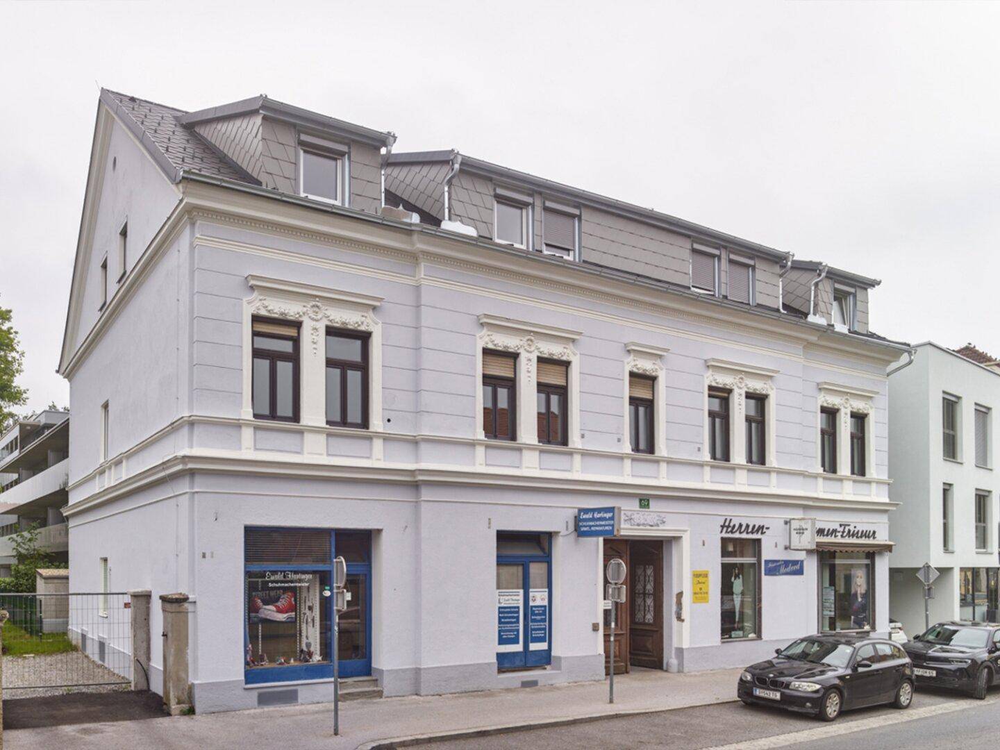 Eckgebäude mit klassischer Fassade, Geschäften im Erdgeschoss und einem modernen Dachausbau.