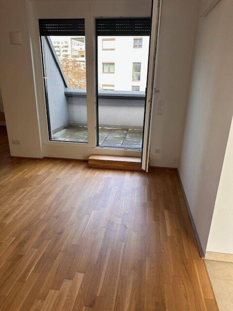 Wohnzimmer mit Holzboden und Zugang zu einem kleinen Balkon mit Blick auf andere Gebäude.