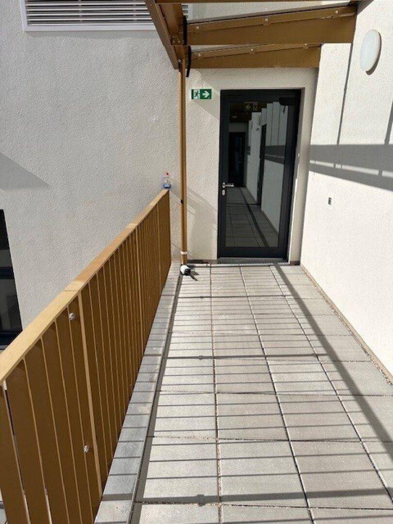 Langer, schmaler Balkon mit Holzboden und Metallgeländer, der zu einem Eingang führt.