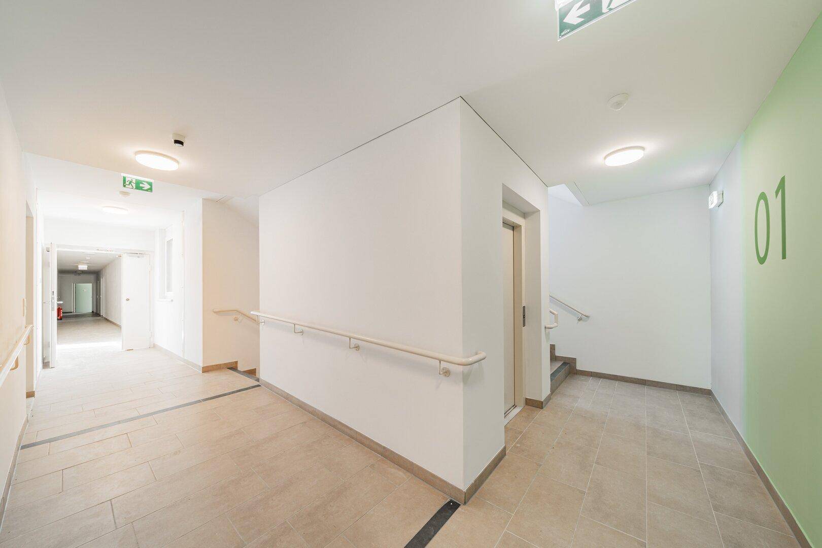Geräumiger Flur im Gebäude mit Aufzug, Treppenhaus und Handläufen.