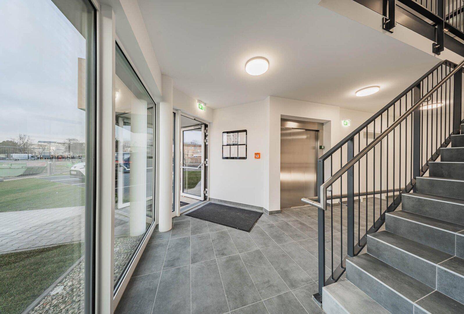 Heller Eingangsbereich mit Treppenhaus, Aufzug und großen Fenstern, die Tageslicht hereinlassen.