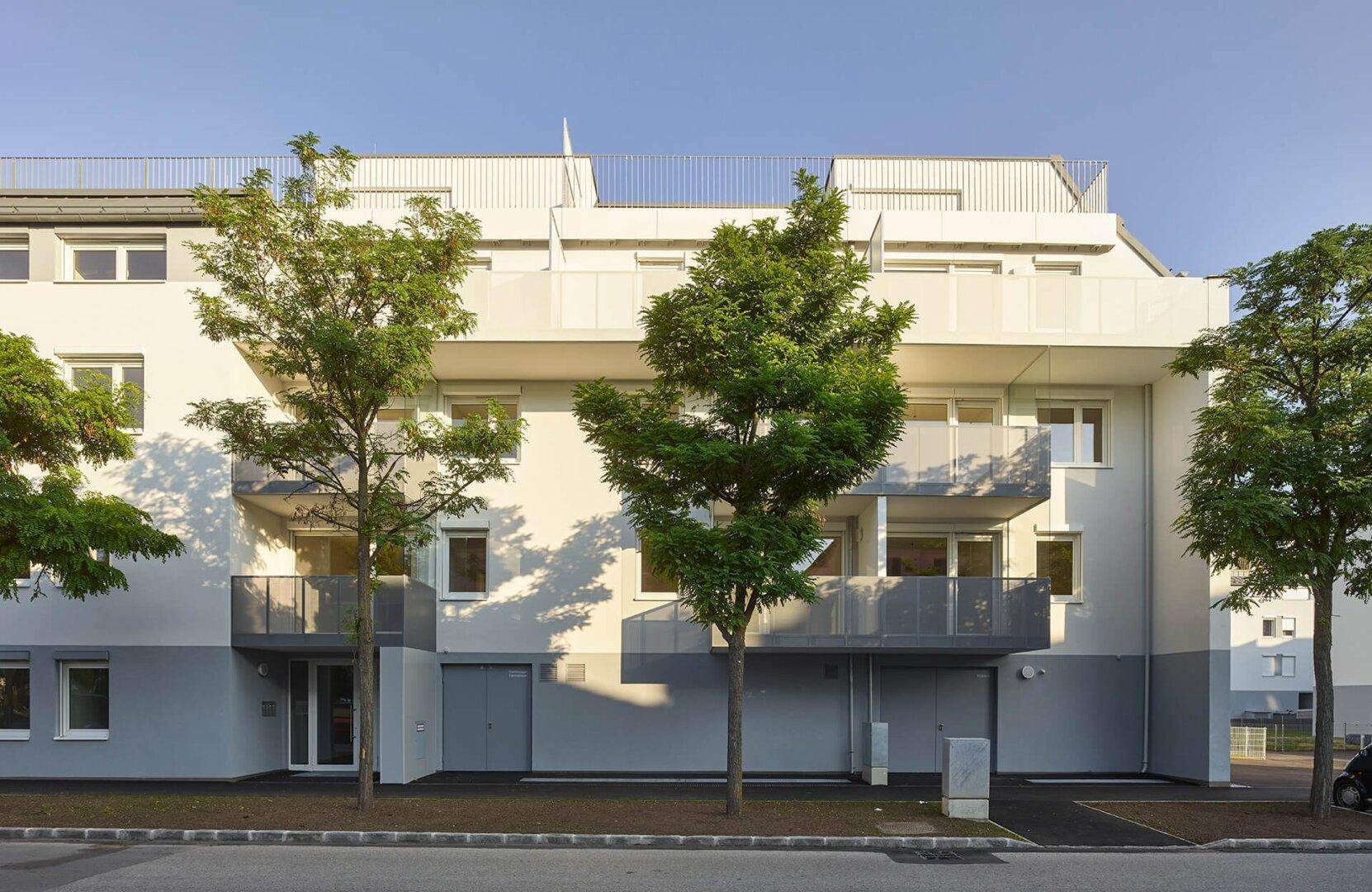Vorderansicht eines Wohnhauses mit Balkonen und Bäumen, die Schatten auf die helle Fassade werfen.