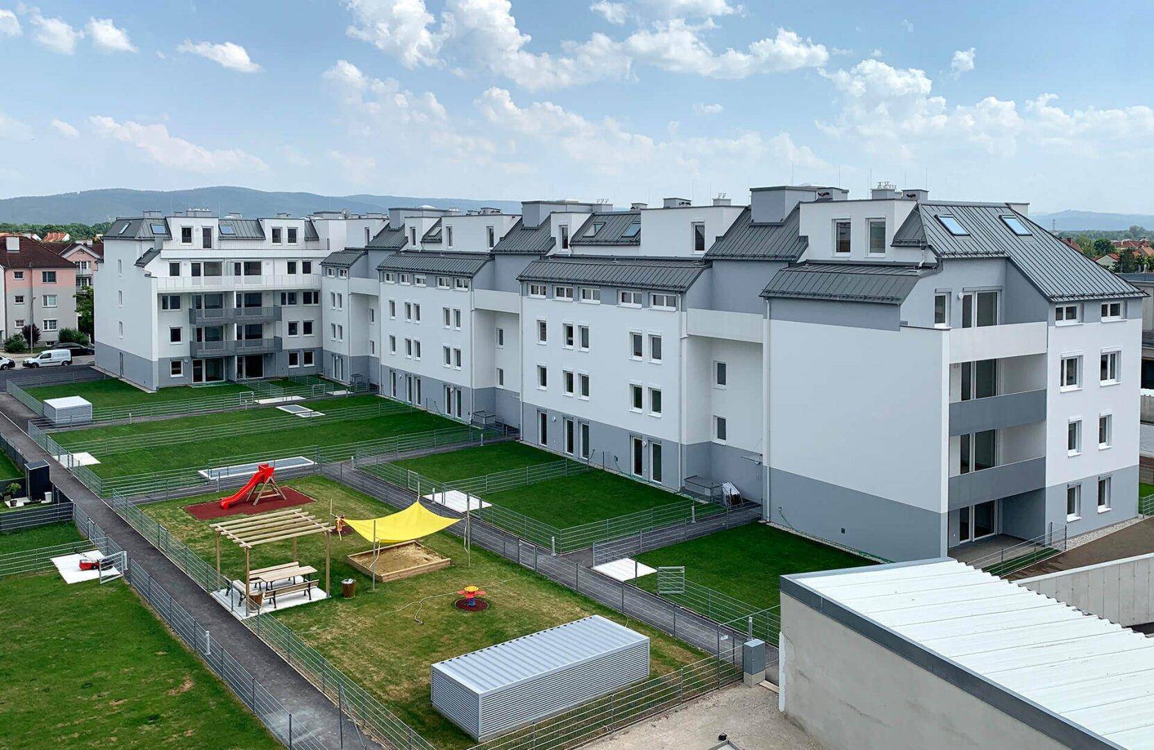 Luftaufnahme einer modernen Wohnanlage mit mehreren Gebäuden, Grünflächen und einem Kinderspielplatz.