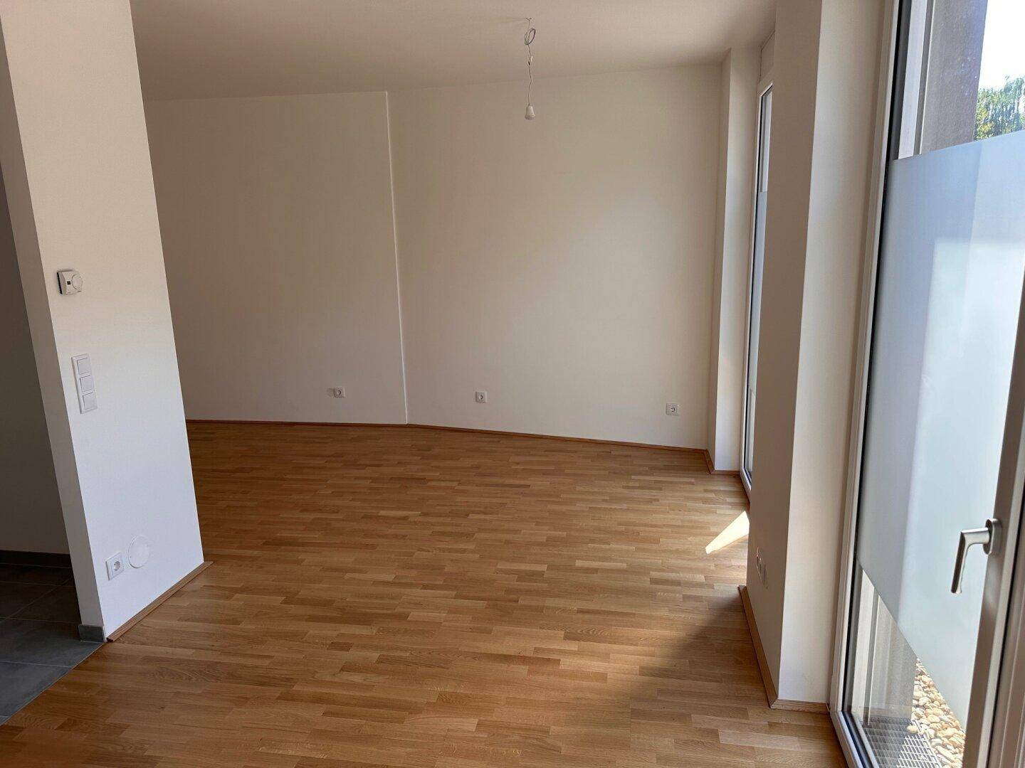 Leerer Wohnraum mit hellem Holzboden und großen Fenstern, die viel Tageslicht hereinlassen.