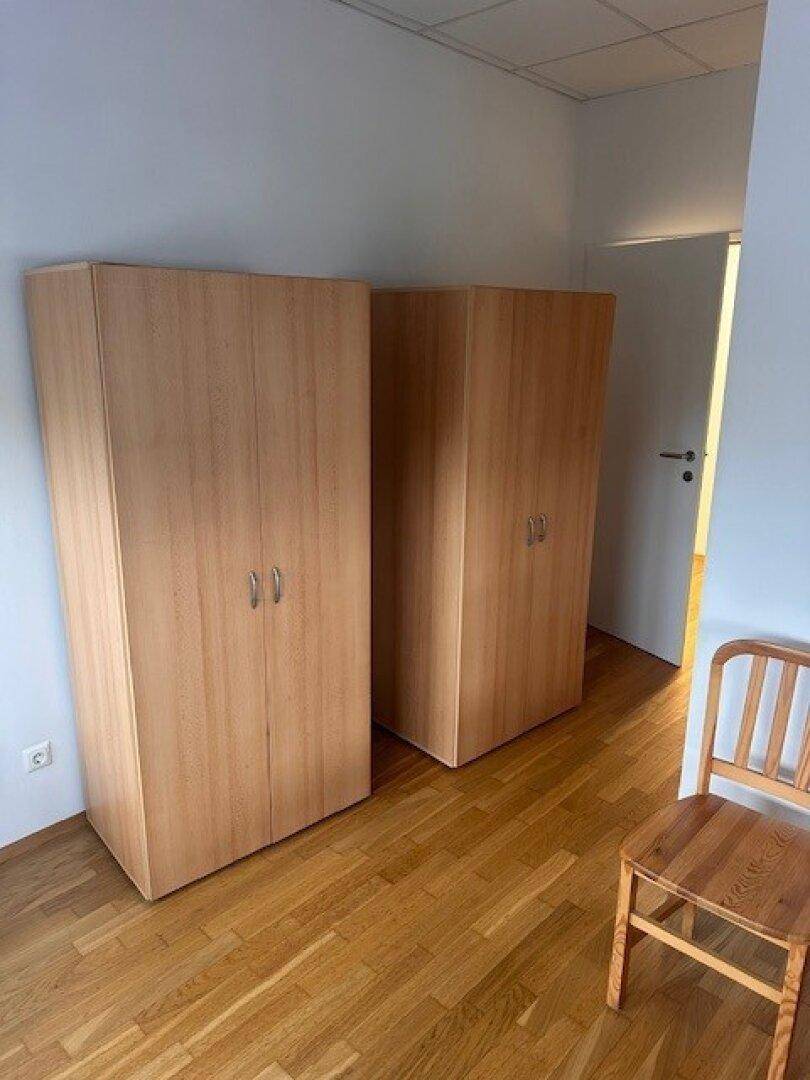 Schlafzimmer mit zwei Holzschränken und einem Stuhl, Holzboden und weiß gestrichenen Wänden.