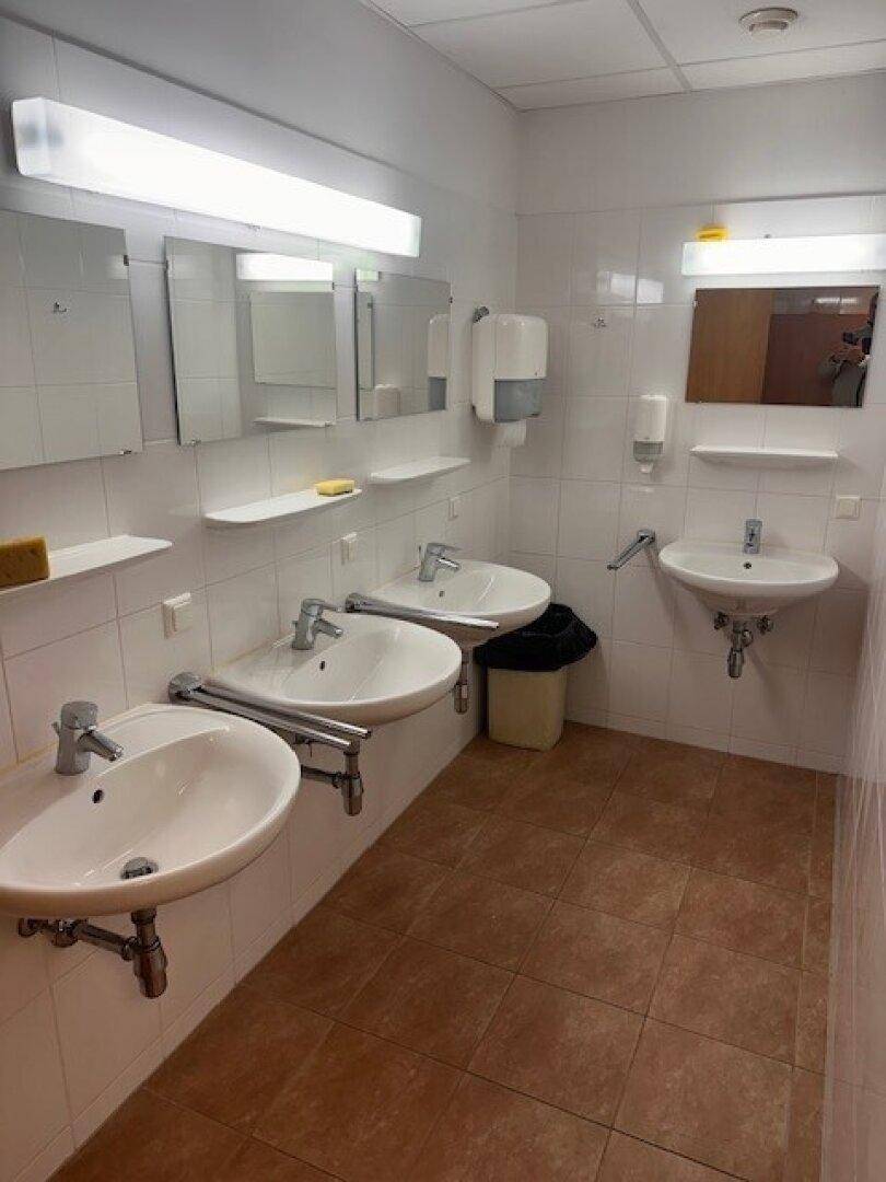 Waschraum mit mehreren Waschbecken, weißen Fliesen und braunem Fliesenboden.