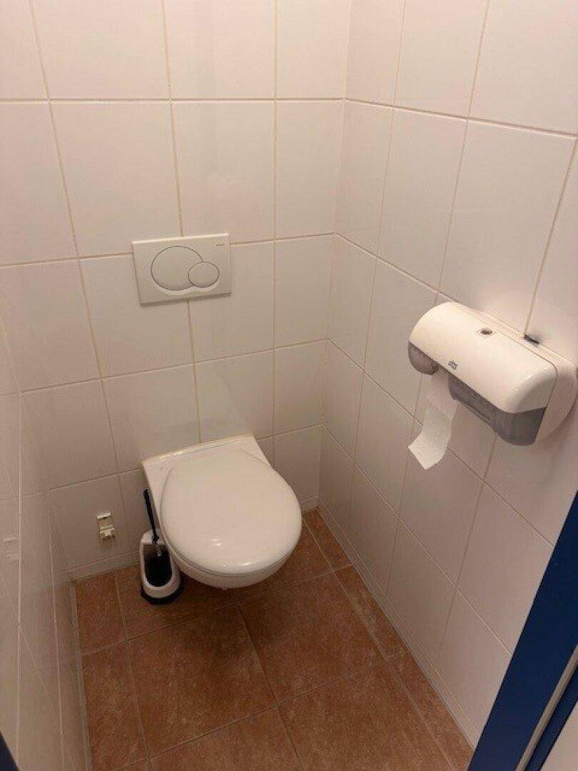Toilettenkabine mit WC, weißen Fliesen und braunem Fliesenboden.