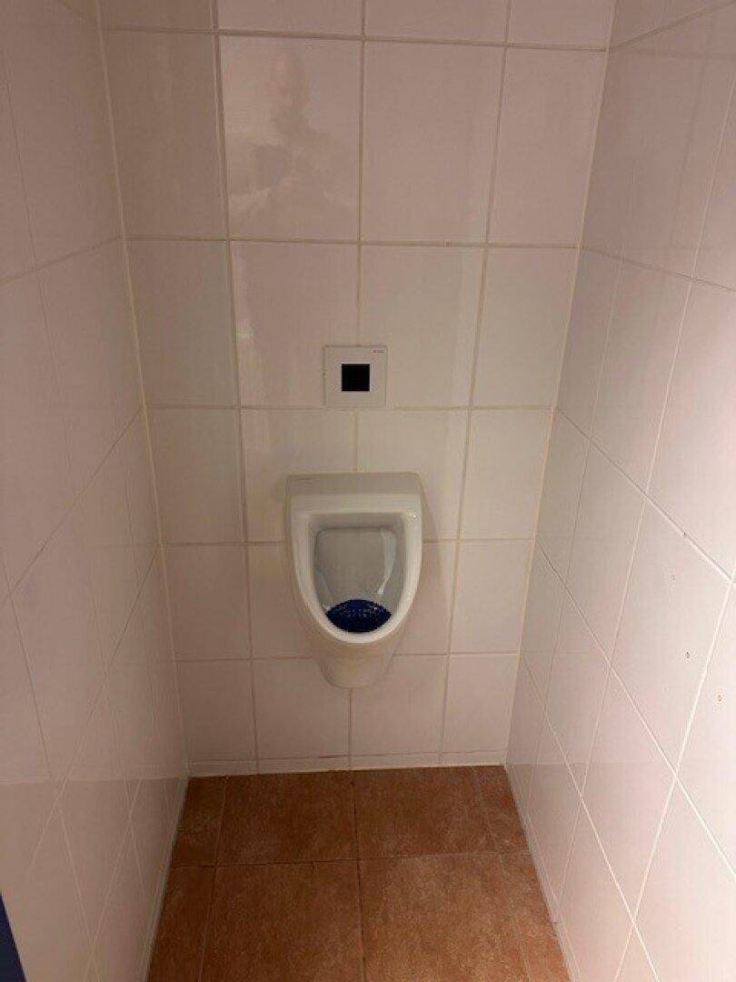 Toilettenkabine mit Urinal, weißen Fliesen und braunem Fliesenboden.