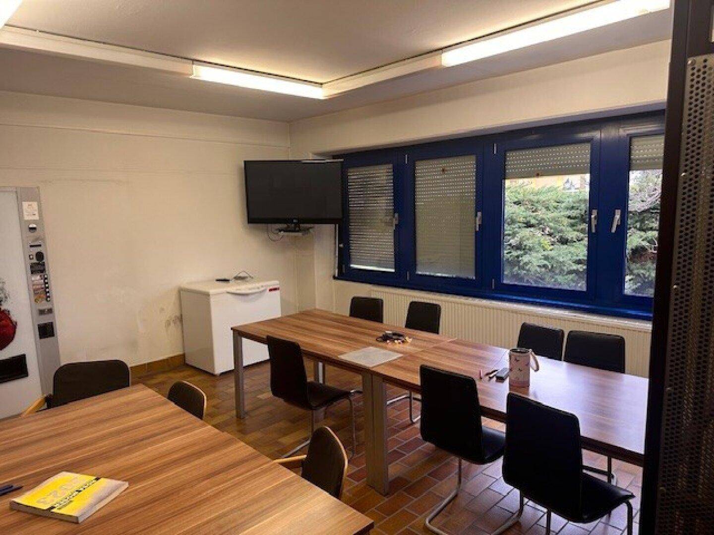 Konferenzraum mit Holztischen, Stühlen und einem Fernseher an der Wand.