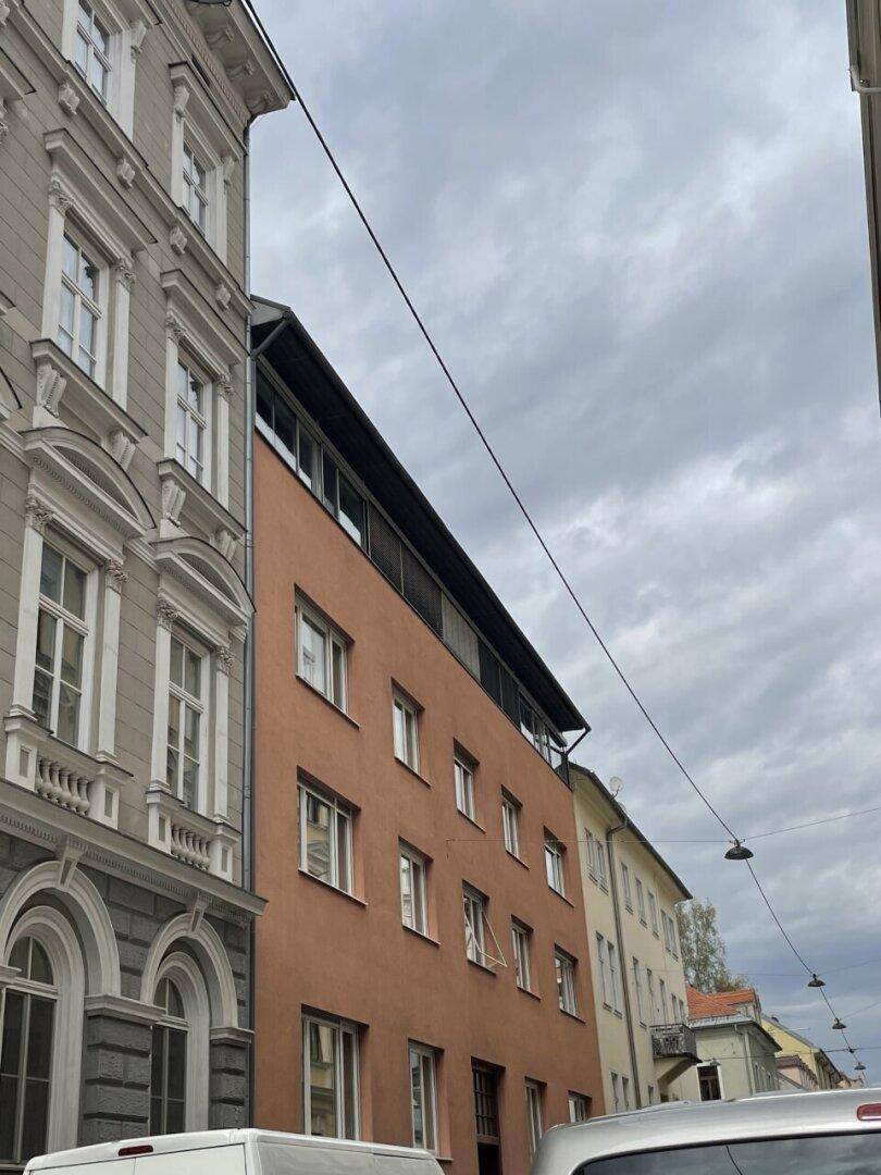 Ansicht einer Stadtstraße mit mehreren Gebäudefassaden, darunter ein modernes Gebäude und ein Altbau, unter bewölktem Himmel.