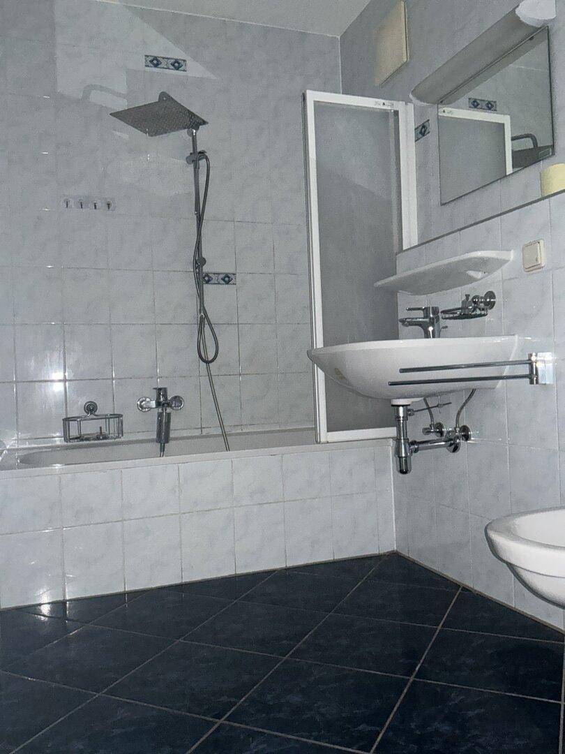 Funktionales Badezimmer mit Badewanne, Duschbereich und hellgrauen Fliesen an Wänden und Boden.