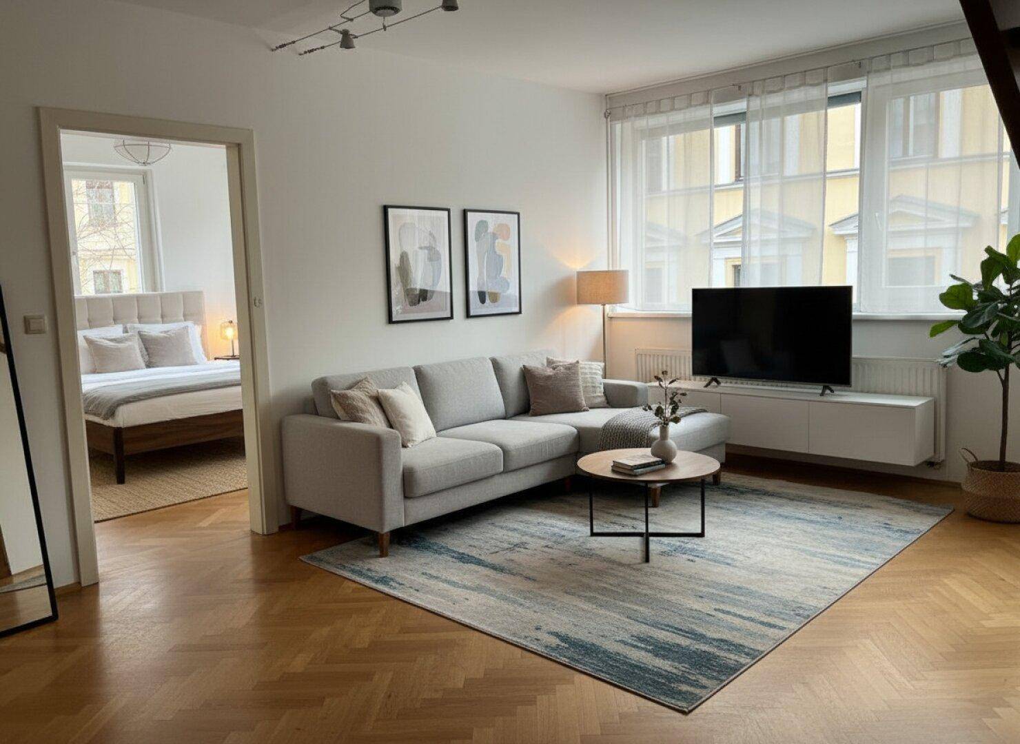 Helles Wohnzimmer mit Parkettboden, bequemer Couch und Blick in ein angrenzendes Schlafzimmer.