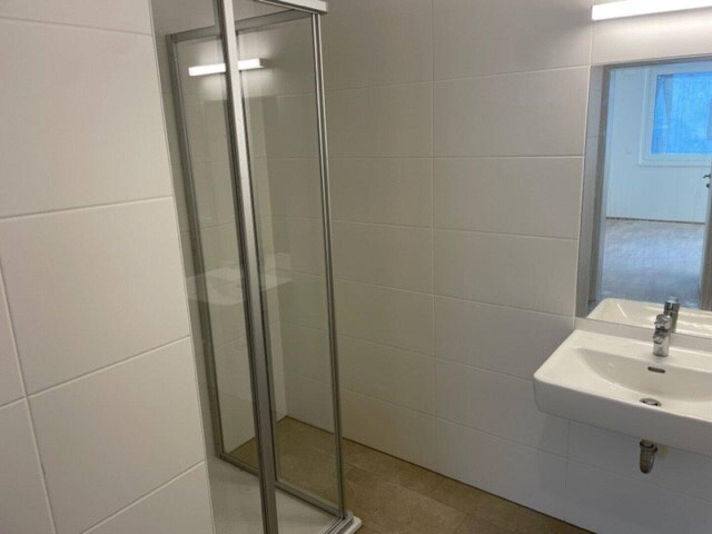 Modernes Badezimmer mit Duschkabine und Waschbecken, durch eine Tür mit einem weiteren Raum verbunden.