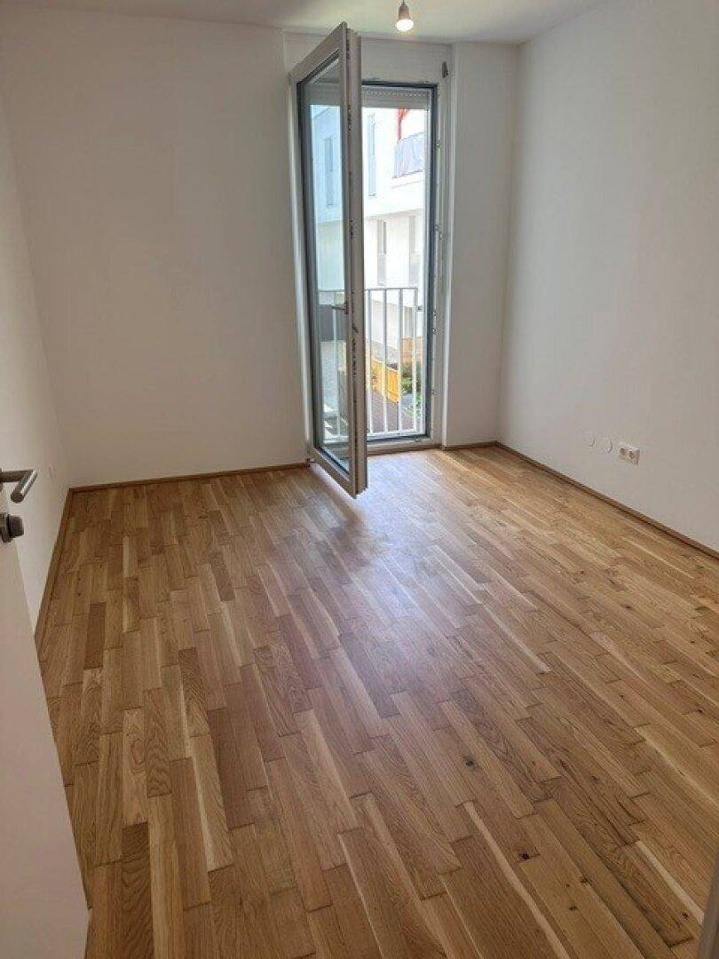Freundliches Zimmer mit Parkettboden und einer Balkontür, die viel Tageslicht hereinlässt.
