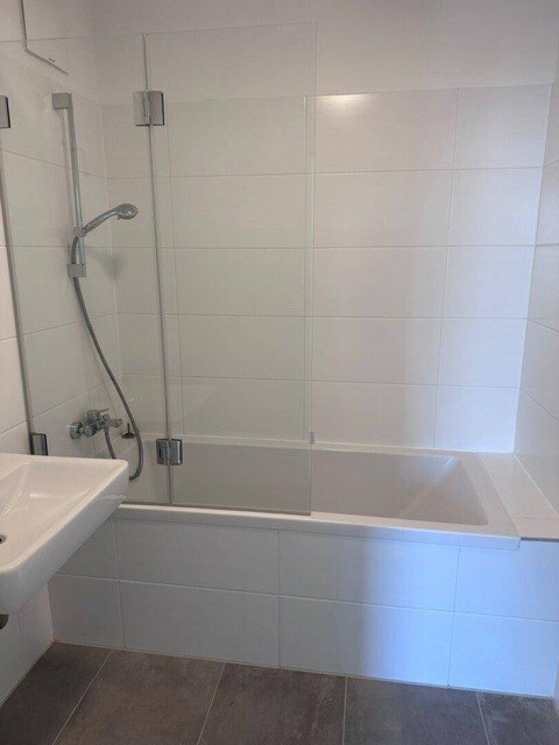 Badezimmer mit integrierter Badewanne und Glasabtrennung, umgeben von weißen Wandfliesen.