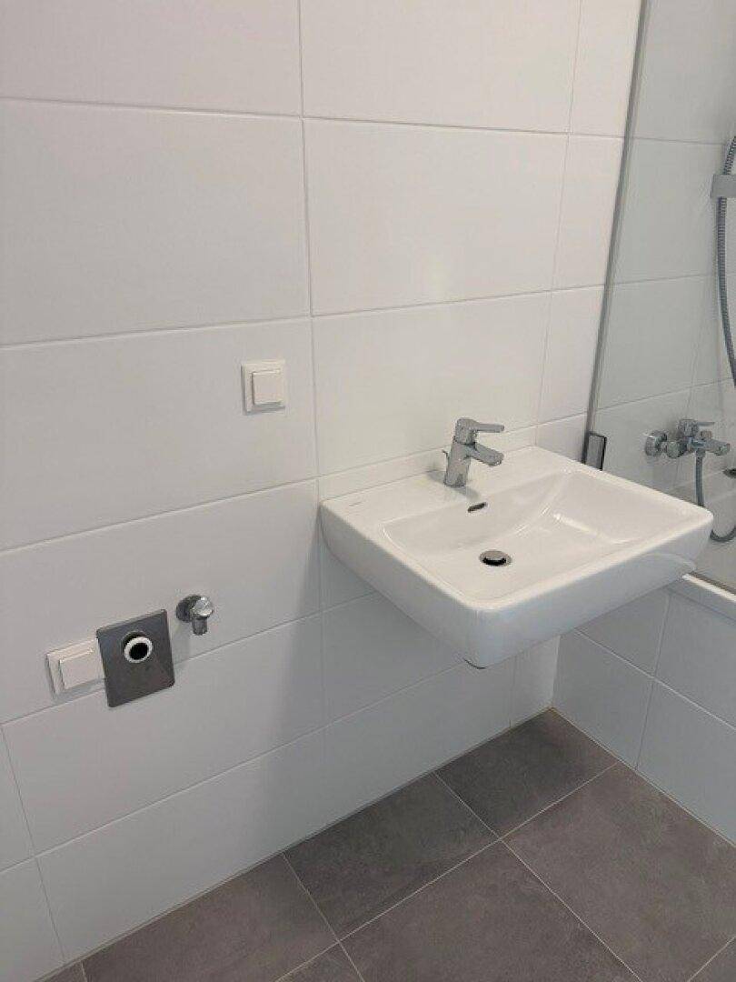 Modernes Badezimmer mit wandmontiertem Waschbecken, weißen Fliesen und grauen Bodenfliesen.
