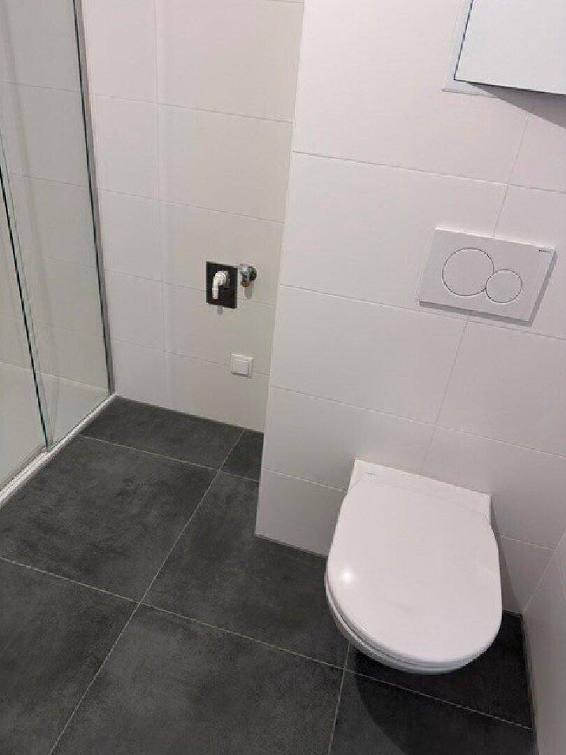 Wandhängende Toilette mit moderner Spülung in einem hellen Badezimmer mit grauen Fliesen.