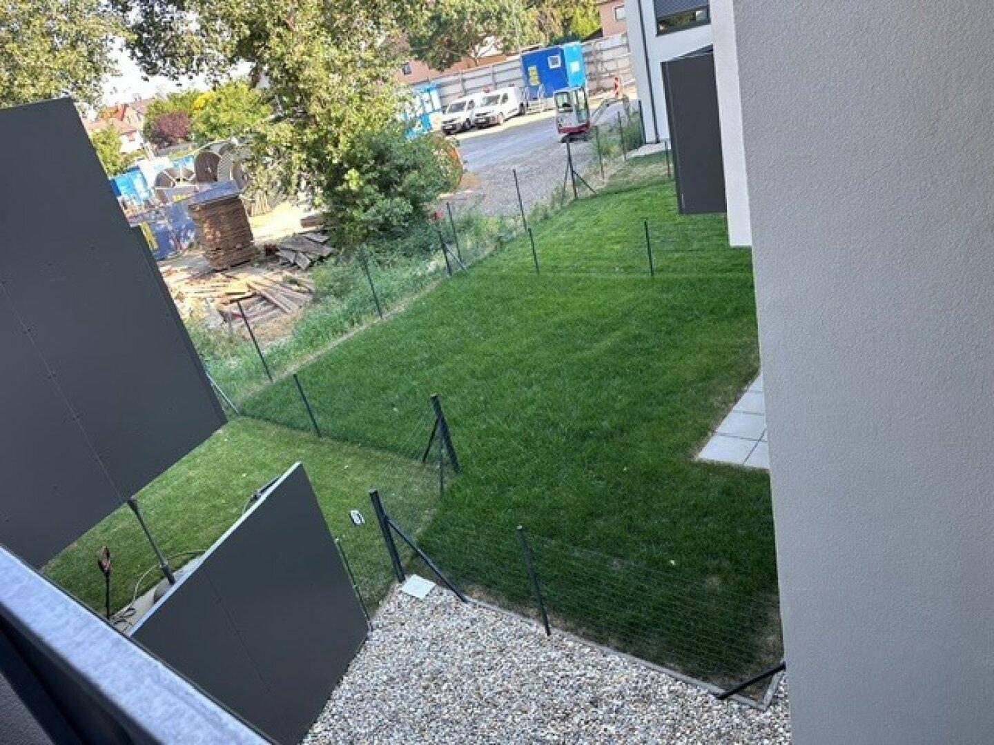 Grüne Rasenfläche, umzäunt, mit Blick auf eine Baustelle im Hintergrund.