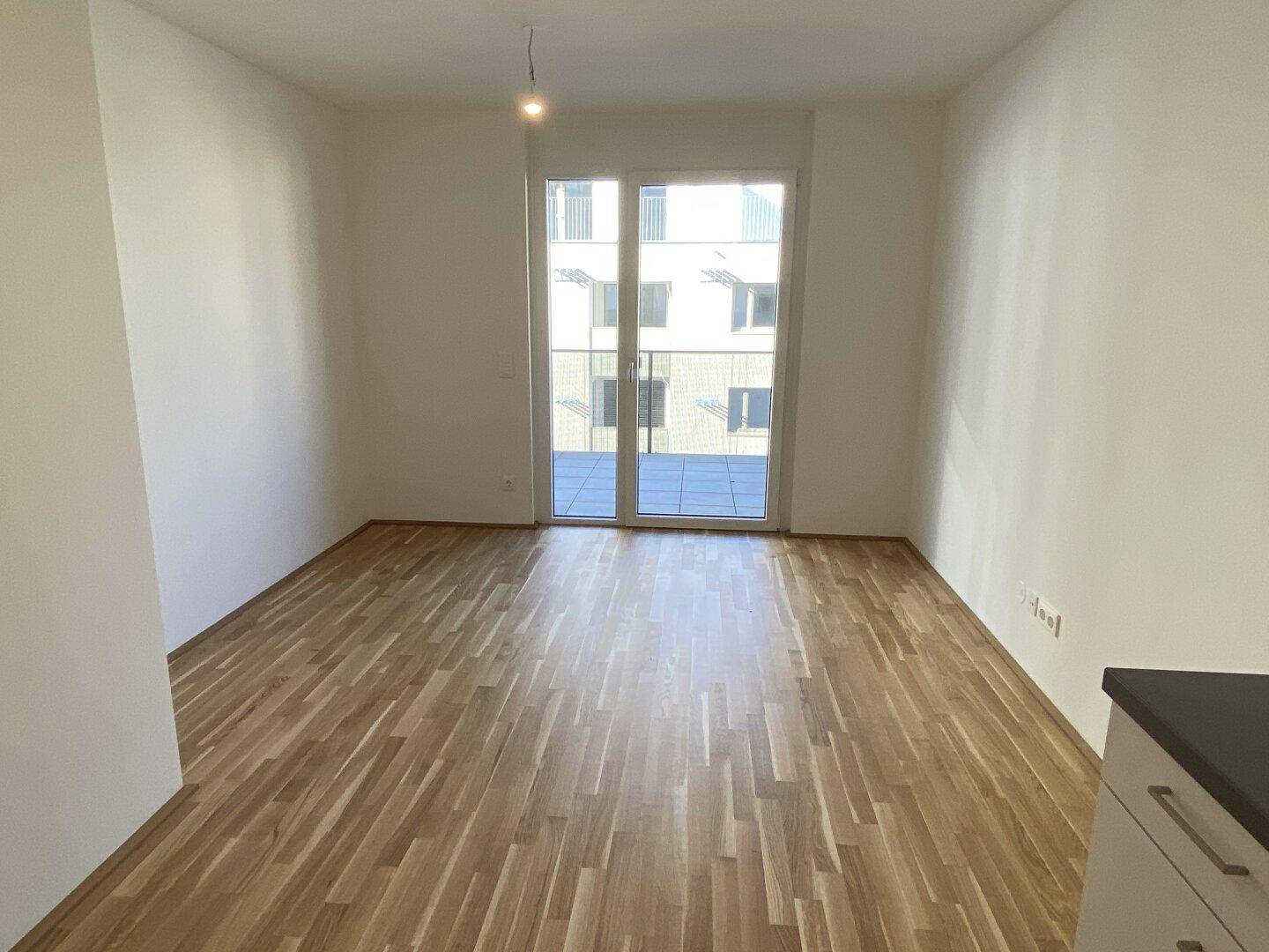 Geräumiges Wohnzimmer mit hellem Holzboden und großer Balkontür, die viel Licht hereinlässt.