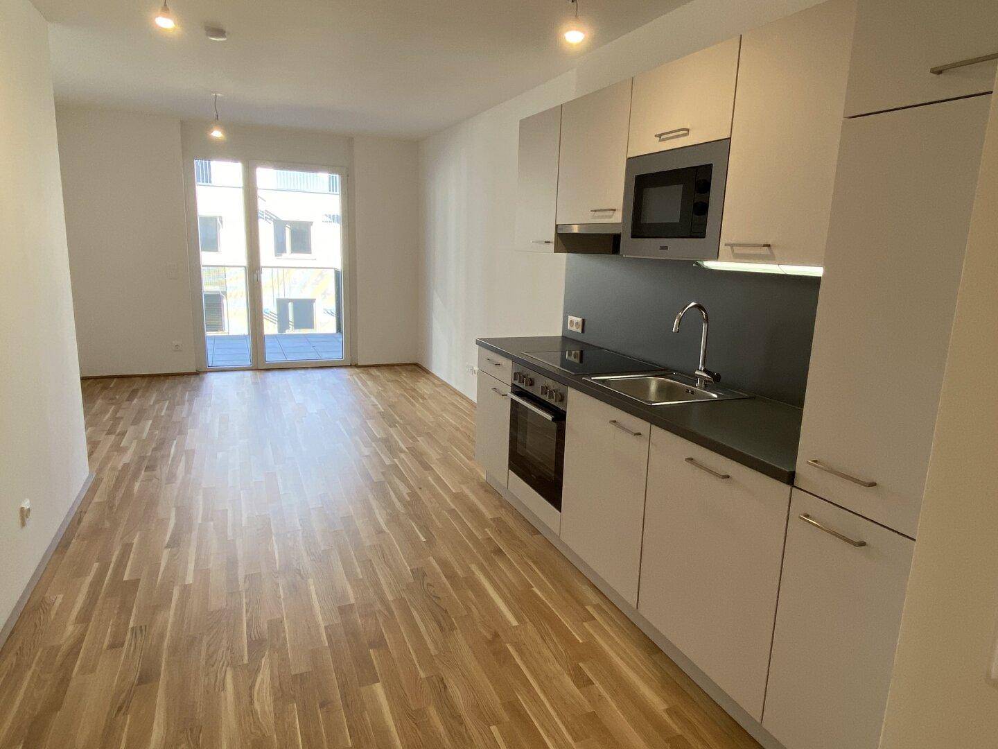 Offener Wohnbereich mit integrierter Küchenzeile, Holzboden und Zugang zum Balkon.