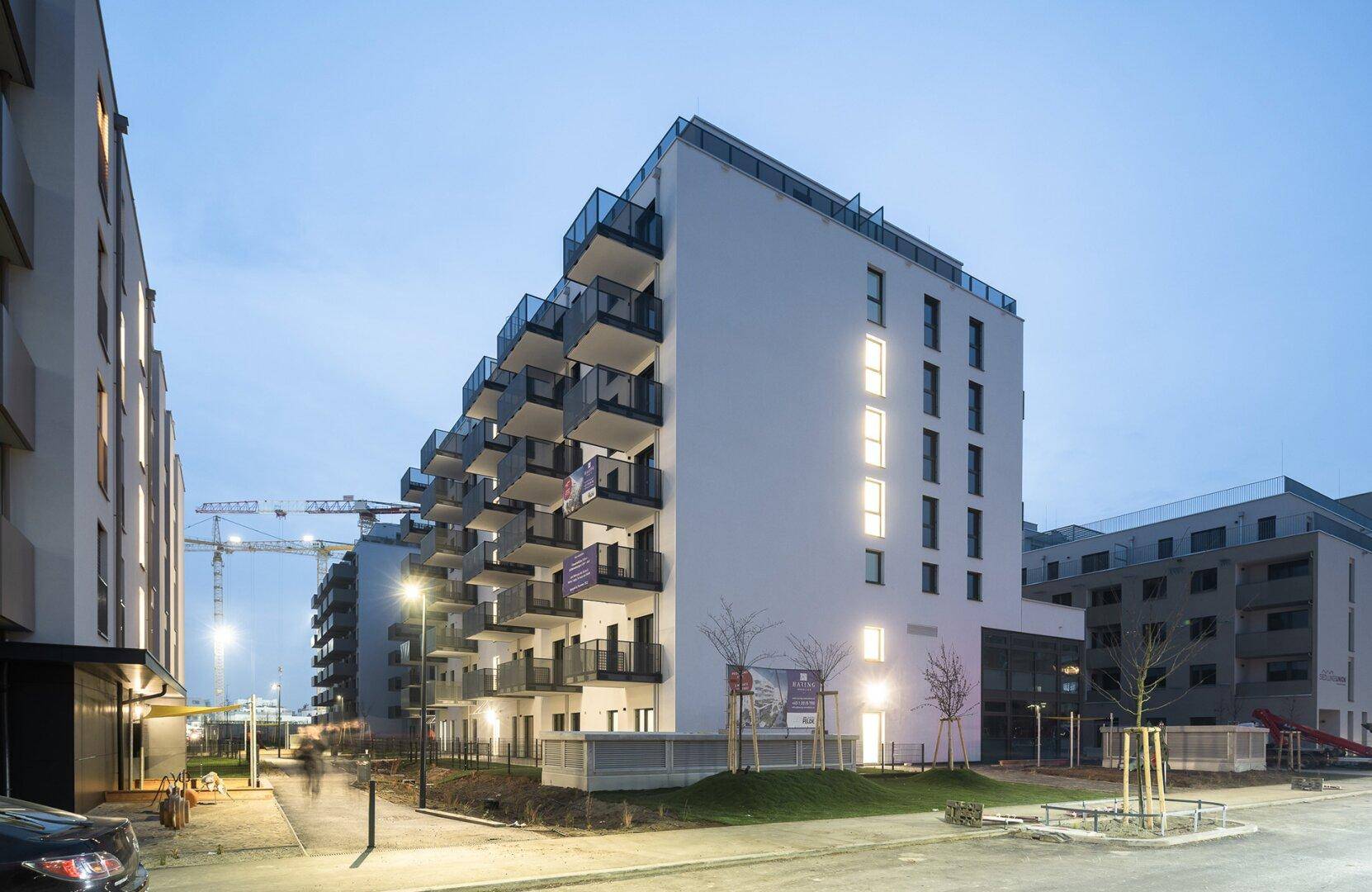 Modernes Wohngebäude bei Dämmerung mit beleuchteten Fenstern und Balkonen in urbaner Umgebung.