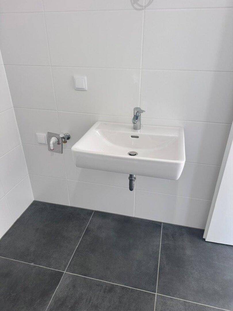 Helles Badezimmer mit weißem Waschbecken und Wandfliesen, ergänzt durch dunkle Bodenfliesen.