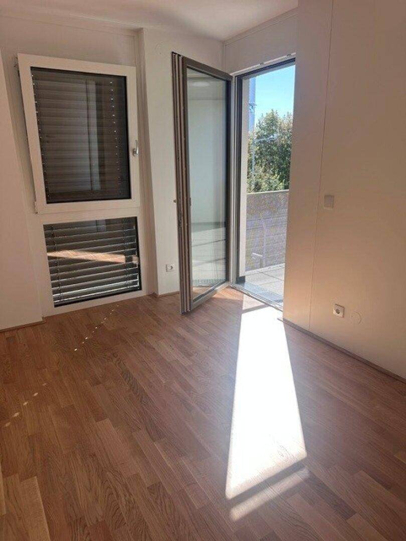 Heller Raum mit Holzboden und Fenster mit Jalousien, bietet Zugang zu einem Balkon.